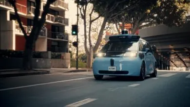 A light blue Waymo autonomous vehicle driving down a sunny city street lined with trees and apartment buildings.