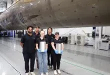 Five aerospace engineers posing in front of a massive rocket component inside a bright manufacturing facility.