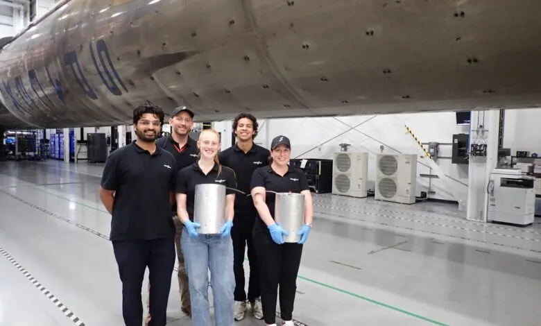 Five aerospace engineers posing in front of a massive rocket component inside a bright manufacturing facility.