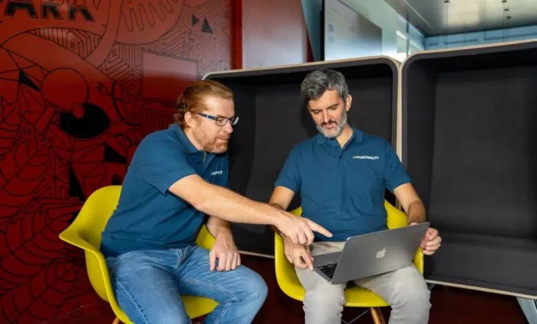 Two men in blue polo shirts collaborating on a laptop in a vibrant modern office space.