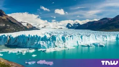 Massive blue glacier wall drops into vibrant turquoise water with dark mountains framing the scene under a blue sky.