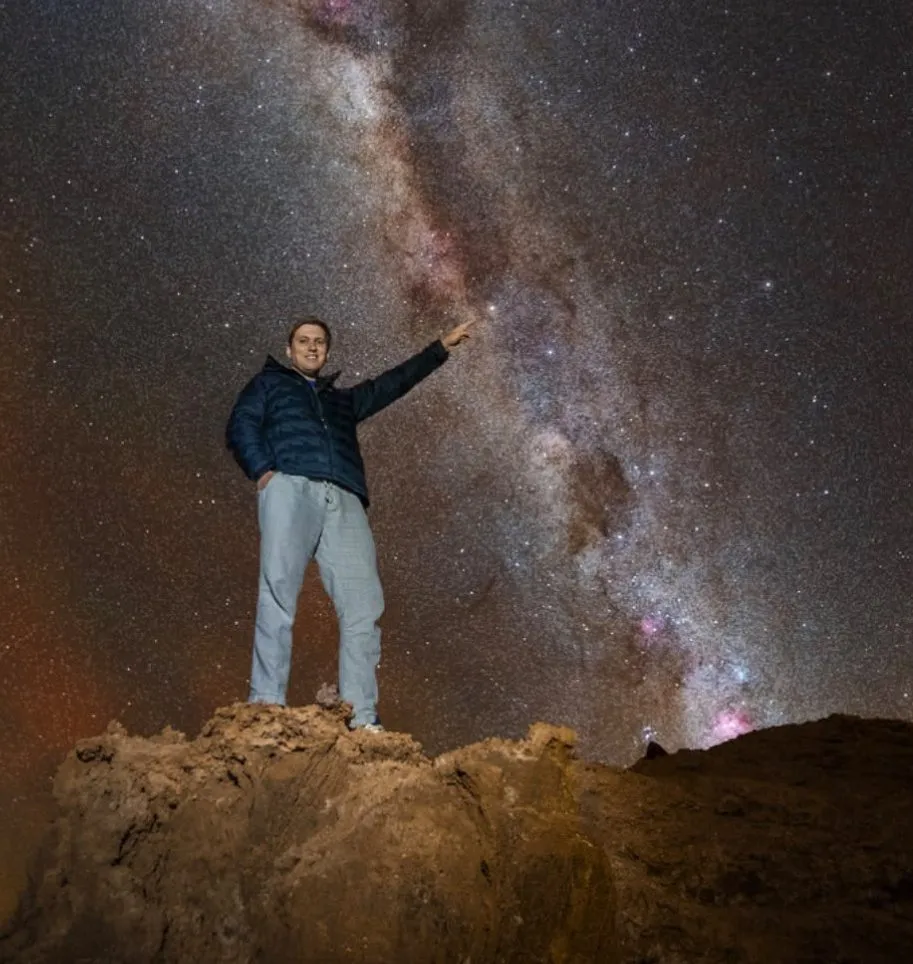 A man stands on a rocky outcrop at night, pointing up at the vast Milky Way galaxy stretching across the starry sky.