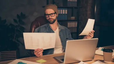 Confused man in a beanie and glasses reviews financial charts and documents at his desk.