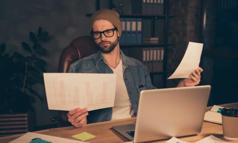 Confused man in a beanie and glasses reviews financial charts and documents at his desk.