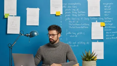 Programmer working at desk with laptop in front of a blue wall with Java code and notes