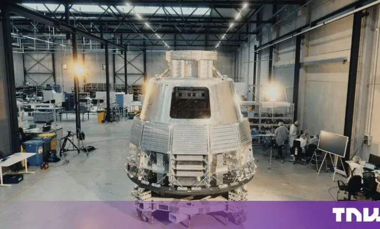 Large metallic space capsule undergoing assembly inside a modern manufacturing warehouse.