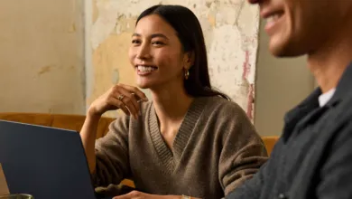 A smiling woman in a cozy brown sweater working on a laptop with a colleague.