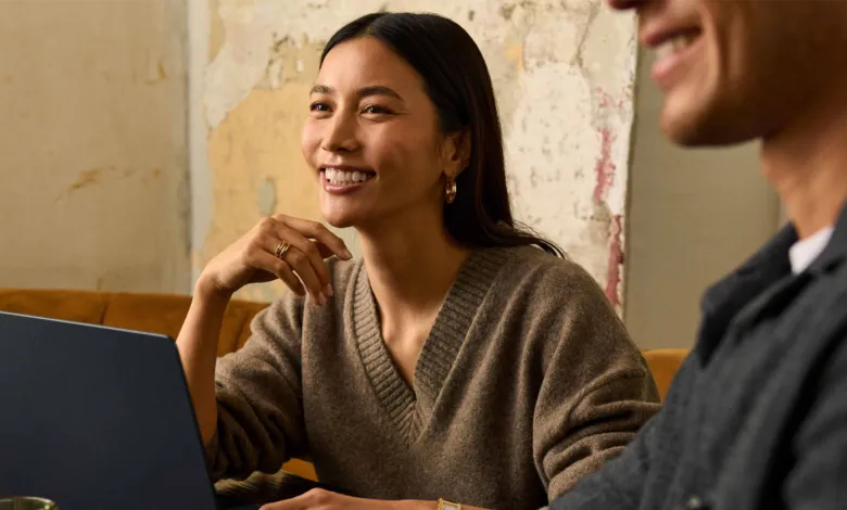 A smiling woman in a cozy brown sweater working on a laptop with a colleague.