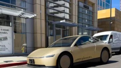 A beige Tesla Cybercab robotaxi driving on a sunny city street past a store under construction.