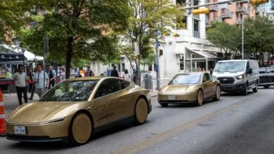 Two gold-wrapped Tesla vehicles driving on a city street past pedestrians and buildings.