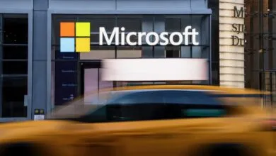 A motion-blurred yellow taxi drives past the modern storefront of a Microsoft office building.