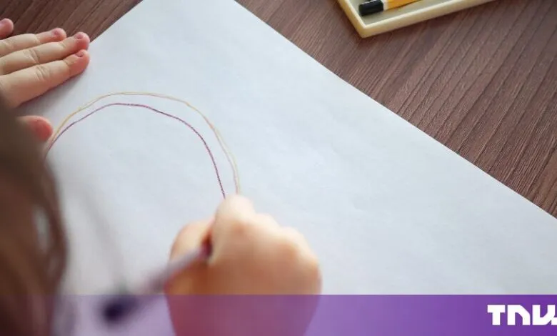 A young child holds a colored pencil, drawing a curved line on a large white sheet of paper.
