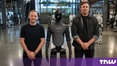 Two men standing next to a black humanoid robot in an indoor facility with other robots in the background.