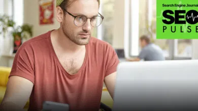 Man wearing round glasses and a maroon t-shirt focuses intensely while working on a laptop computer.
