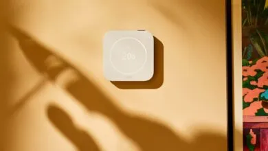 Modern white smart thermostat mounted on a warm beige wall with soft leaf shadows.