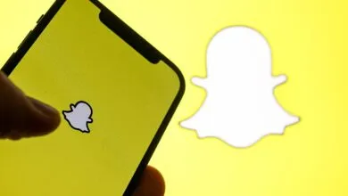 A hand taps the Snapchat app icon on a smartphone with a vibrant yellow background.