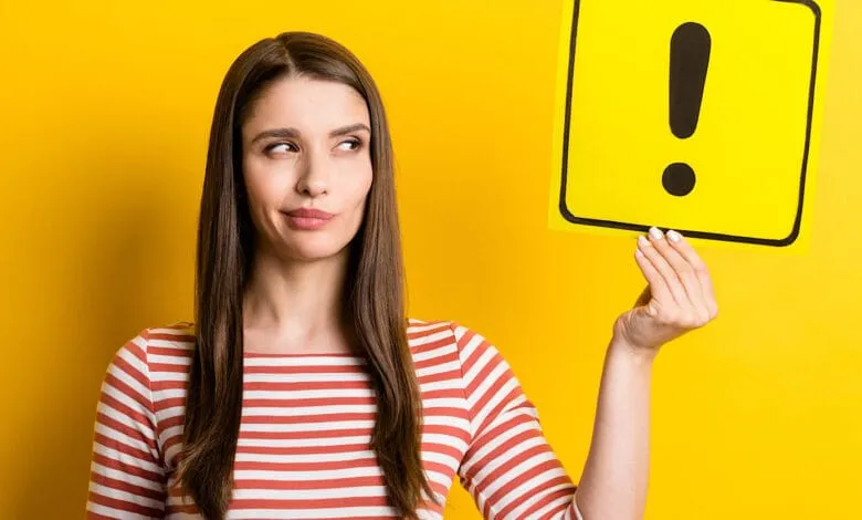 A young woman with long brown hair, wearing a red and white striped shirt, holds up a yellow card with a black exclamation point, looking thoughtful.