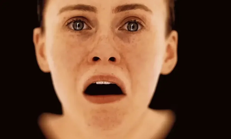 Close-up of a woman's face, eyes wide with fear, mouth open in a gasp against a dark background.