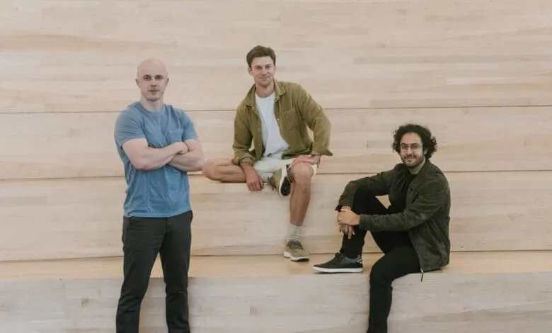 Three men stand and sit against a light wood wall. One stands with arms crossed, two sit with legs crossed.