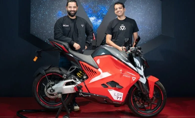 Two men stand proudly beside a sleek, red electric motorcycle. The bike features a futuristic design.