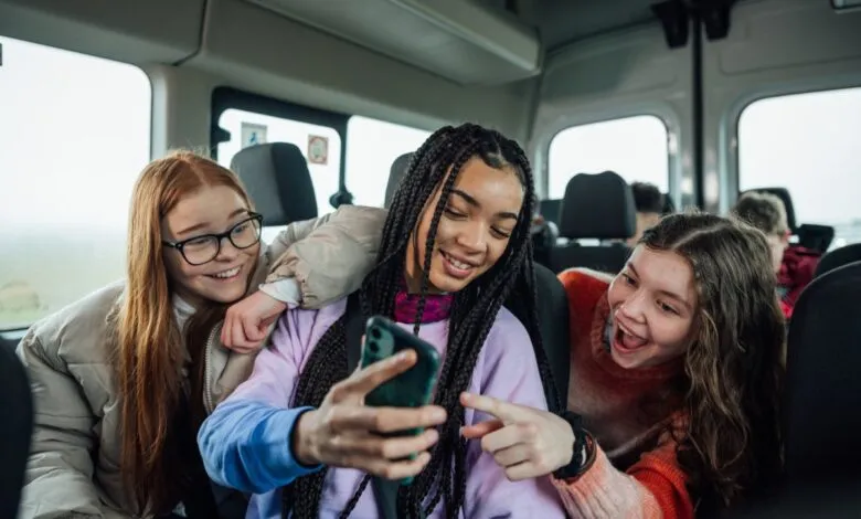 Three teenage girls laughing while looking at a smartphone in a vehicle.