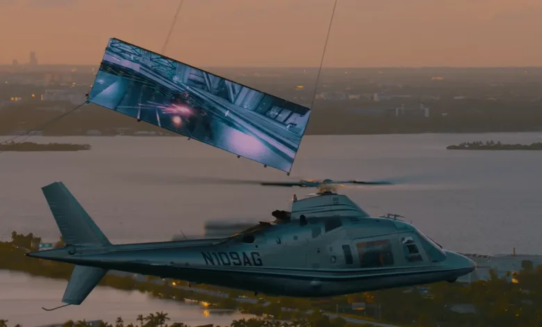 Helicopter carrying a large screen displaying a video game scene over a city at sunset.