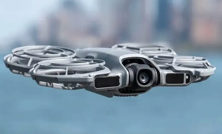 A compact, gray DJI Mini drone with its camera facing forward, hovering against a blurred blue sky.