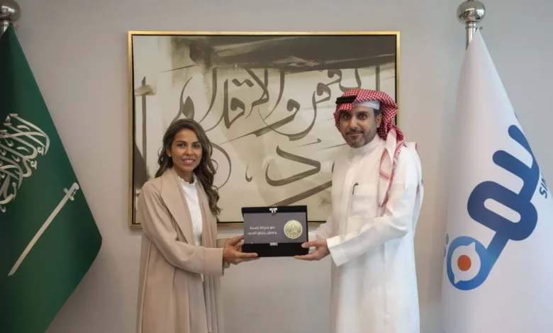 A man and woman exchange a framed award in an office with flags.