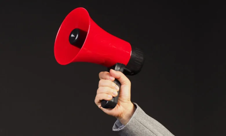A hand in a grey suit jacket holds a bright red megaphone against a dark background.