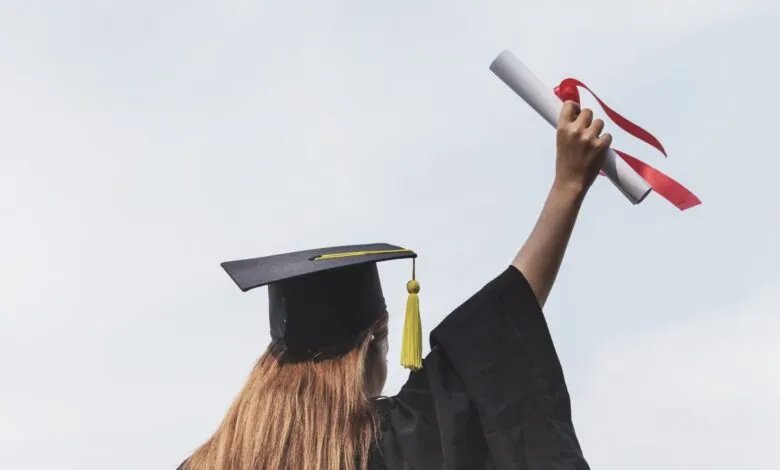 Graduate in cap and gown holds diploma high against sky.