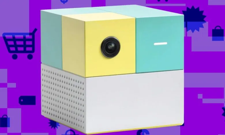 Colorful cube-shaped device with camera lens on yellow face, against a purple shopping-themed background.