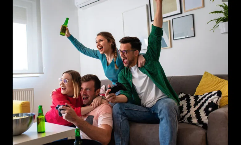 Four friends celebrate while playing video games, holding controllers and beers.