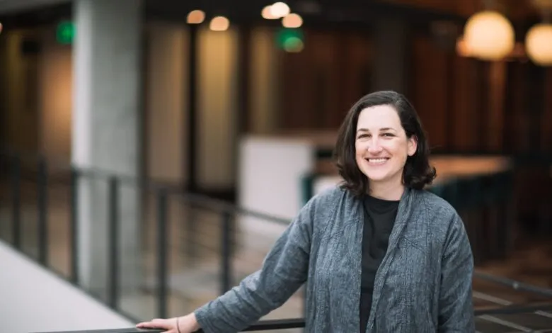 Smiling woman with dark hair stands indoors by a railing.