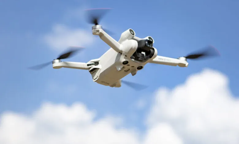 White DJI Mini 3 Pro drone flying against a blue sky with fluffy white clouds.
