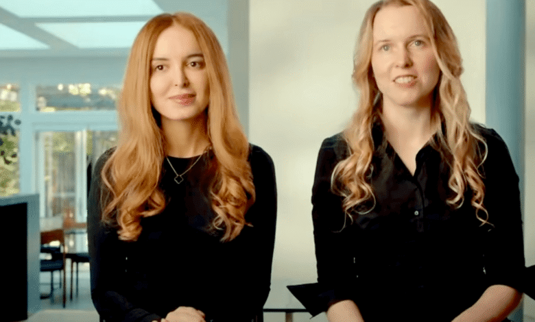 Two women with long hair sit side-by-side in black shirts and jeans, smiling slightly.