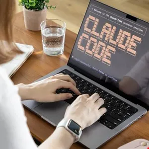 Person types on laptop displaying 'Claude Code' welcome screen, with glass of water nearby.