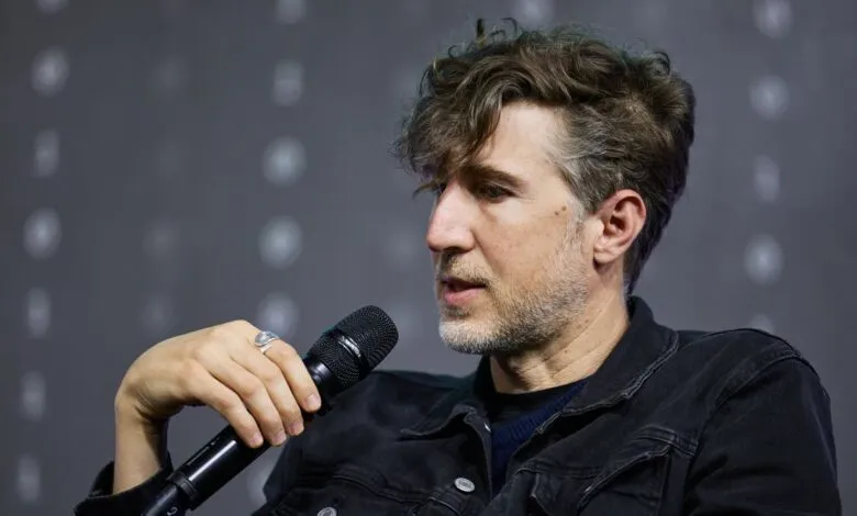 Man with curly hair holds microphone, wearing denim jacket.
