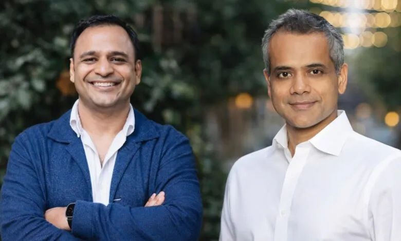 Two men smiling, one in a blue jacket, the other in a white shirt, against a blurred background.