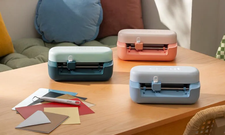 Three colorful Cricut Joy machines rest on a wooden table with crafting supplies.