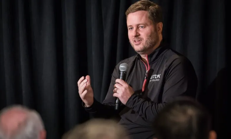 Man speaks into microphone at event, gesturing with hand.