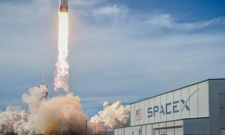 SpaceX rocket launches with bright flames and smoke against a blue sky.
