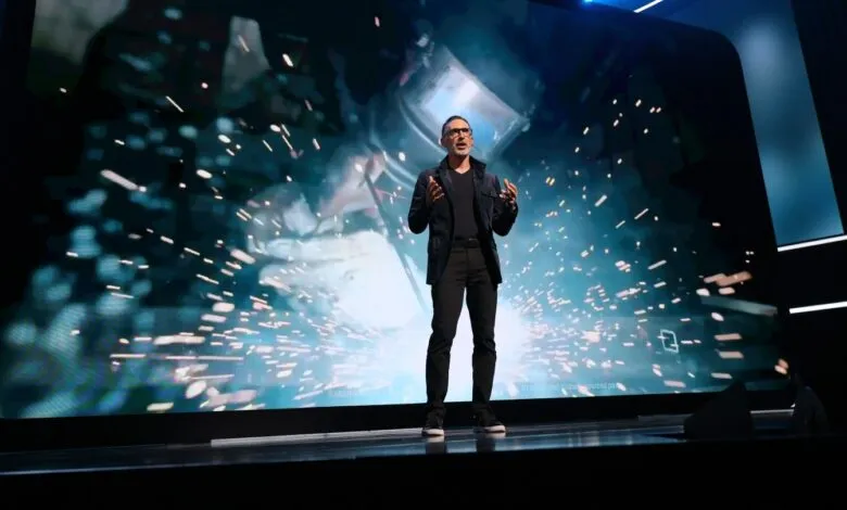 Man on stage presenting with welding sparks on large screen behind him.