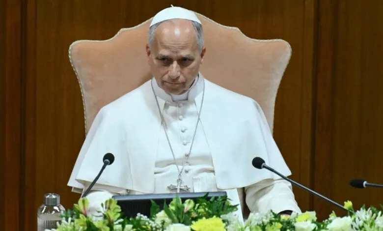 Pope Leo XIV seated at a table with microphones and floral arrangements.