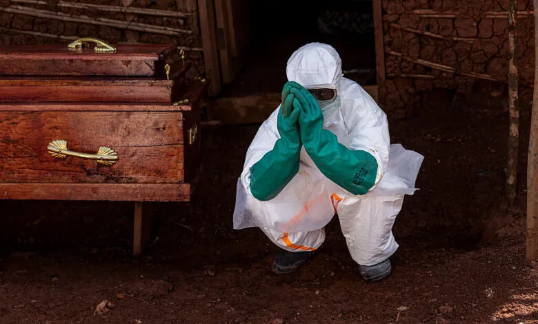 Health worker in PPE crouches near coffin, hands clasped in prayer.