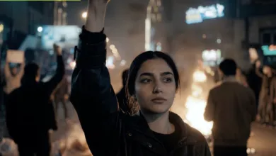 Woman with raised fist at protest, fire burning in background, determined expression.
