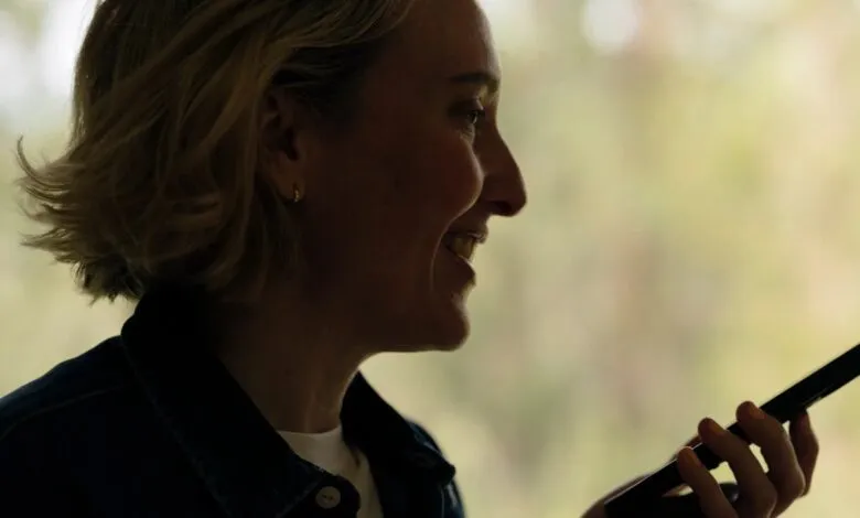 Smiling woman with short blonde hair holds a smartphone, silhouetted against a bright window.