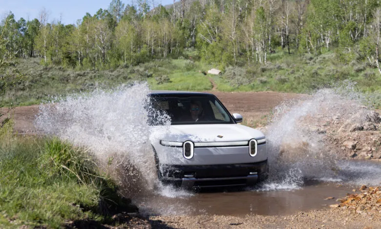 White Rivian electric SUV driving through a shallow stream, splashing water on both sides.