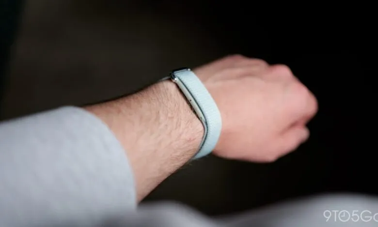 A person wears a light blue Fitbit fitness tracker on their wrist against a dark background.