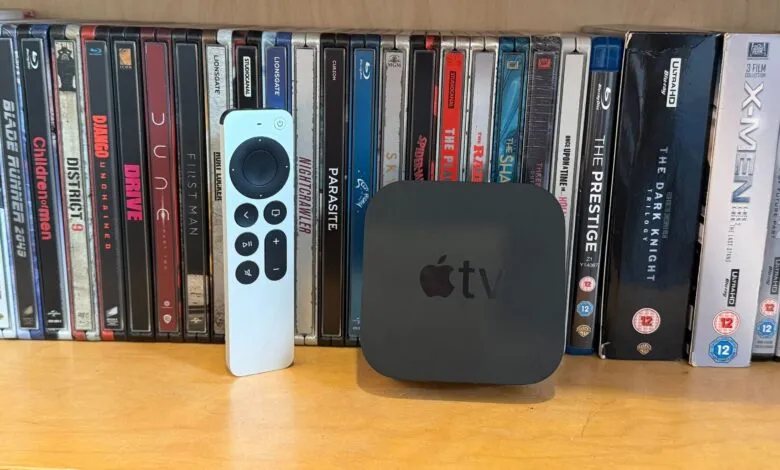 Apple TV box and silver remote resting on a wooden surface against a backdrop of organized steelbook movies.