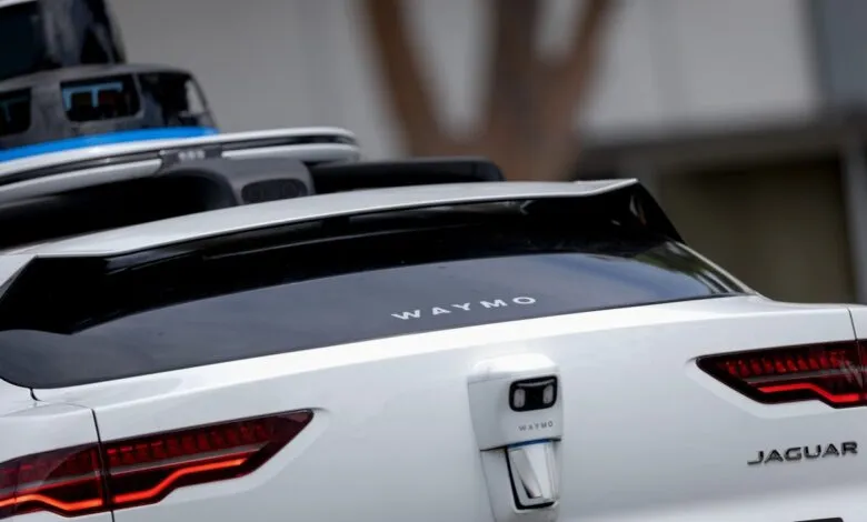 Rear view of a white Waymo autonomous Jaguar vehicle with roof-mounted sensors and Waymo branding.